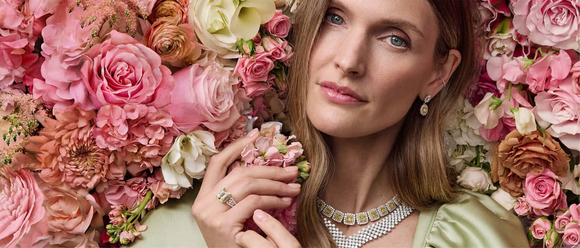 Woman surrounded by pink and white flowers wearing diamond jewelry and a green dress.