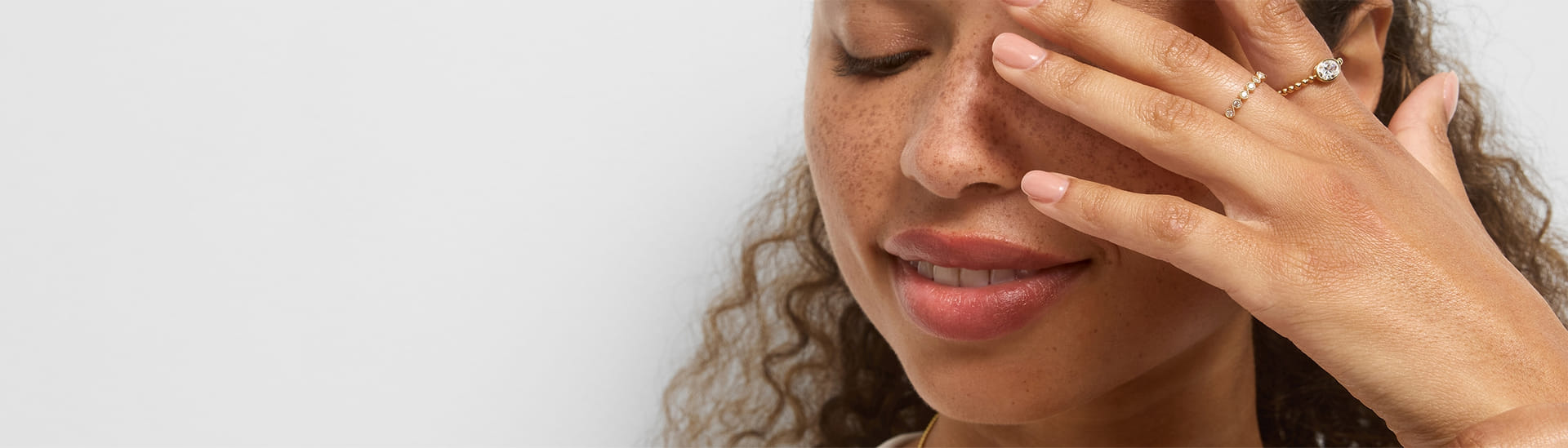 Person with curly hair smiling and covering one eye, wearing gold rings.