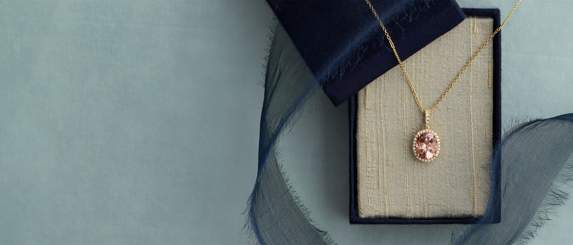 Elegant gold necklace with pink gemstone in a blue-lined box on a soft fabric backdrop.