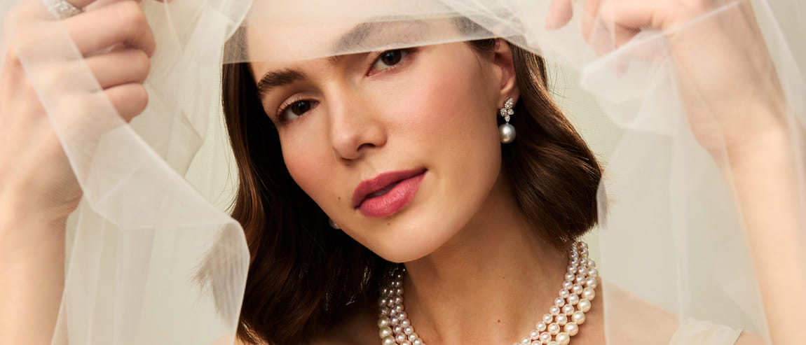 Woman in pearl jewelry holding a veil close to her face.