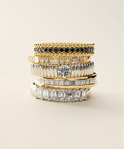 Stack of gold and silver rings with various gemstones and textures on a neutral background.