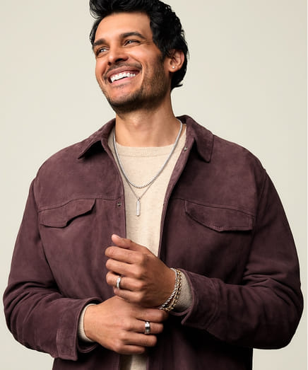 Smiling person wearing a brown jacket with necklaces and rings.