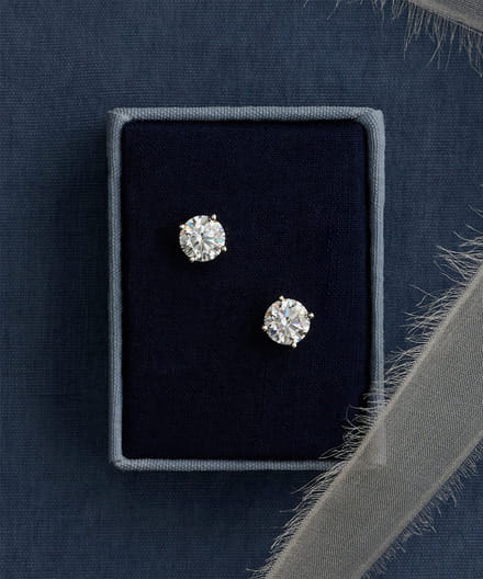 Pair of diamond stud earrings in a dark fabric box with feathered ribbon.