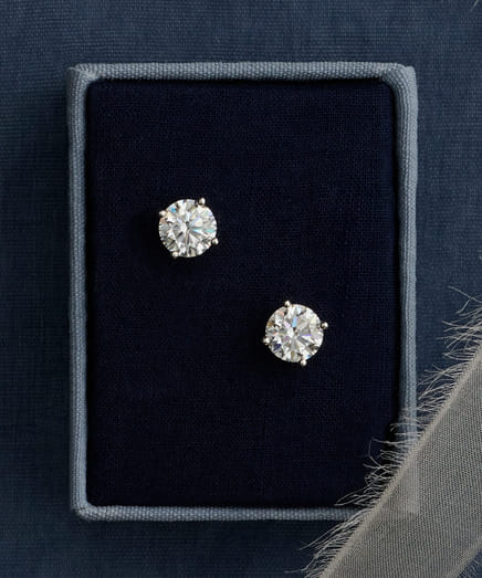 Elegant diamond stud earrings in a jewelry box on a blue fabric background.