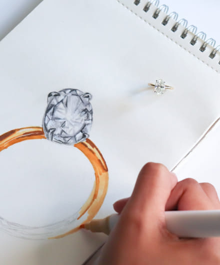 Person sketching a ring design with a gemstone next to a finished ring.