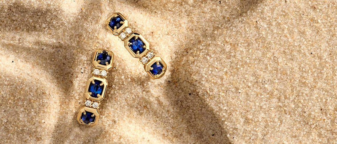 Gold and sapphire drop earrings on textured sand background.