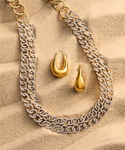 Gold necklace and earrings arranged on a sandy surface.