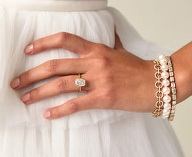 Hand with gold ring and pearl bracelets on white textured fabric.