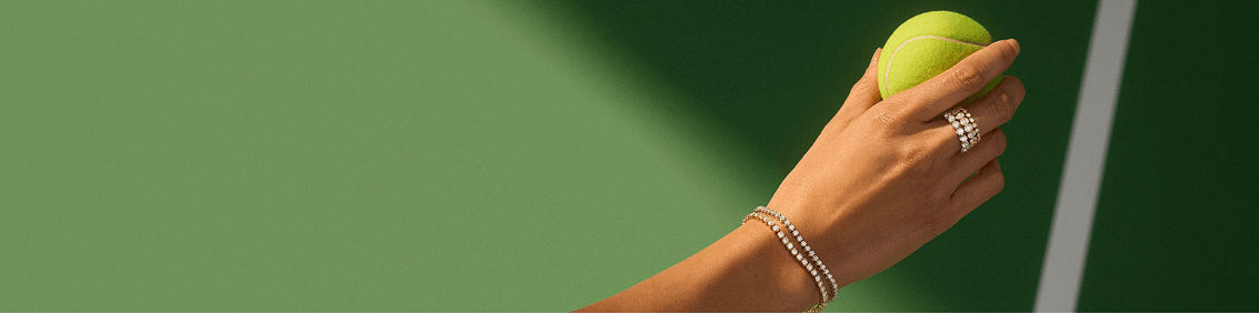 Hand holding a tennis ball adorned with diamond jewelry on a tennis court.