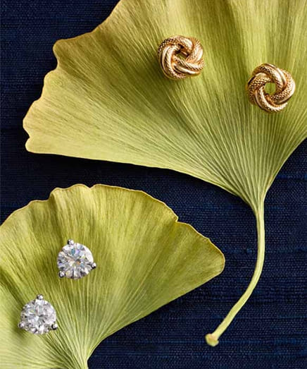 Gold knot and diamond earrings on ginkgo leaves over a dark background.