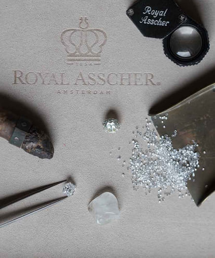 Royal Asscher diamonds with magnifier, tweezers, and rough stone on a branded surface.